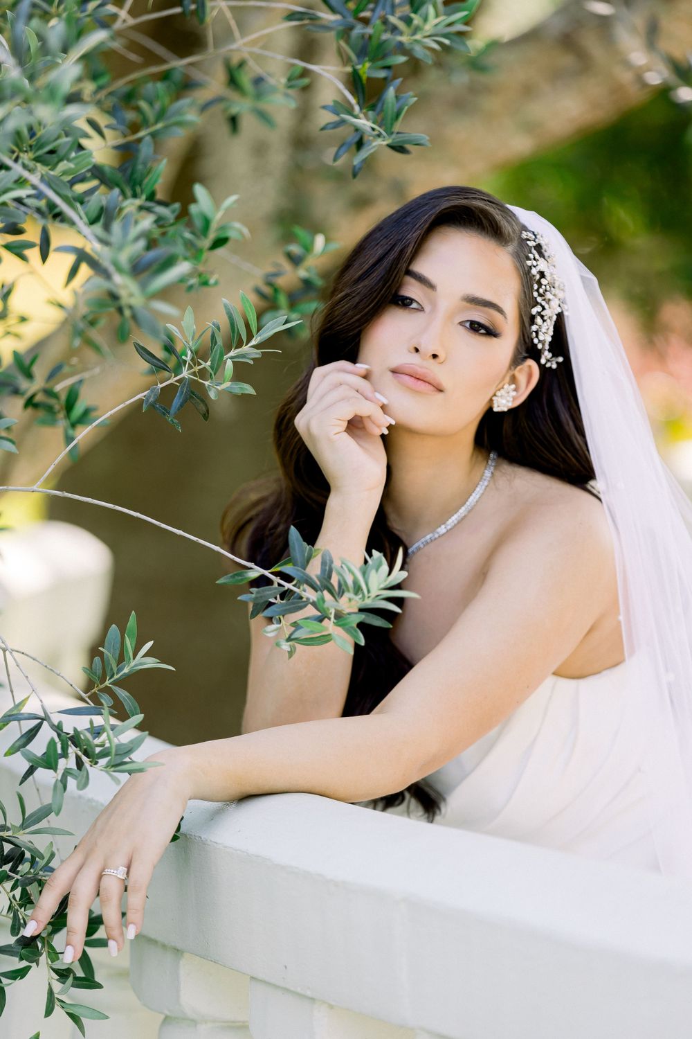 Bride in delicate veil poses thoughtfully among olive branches.
