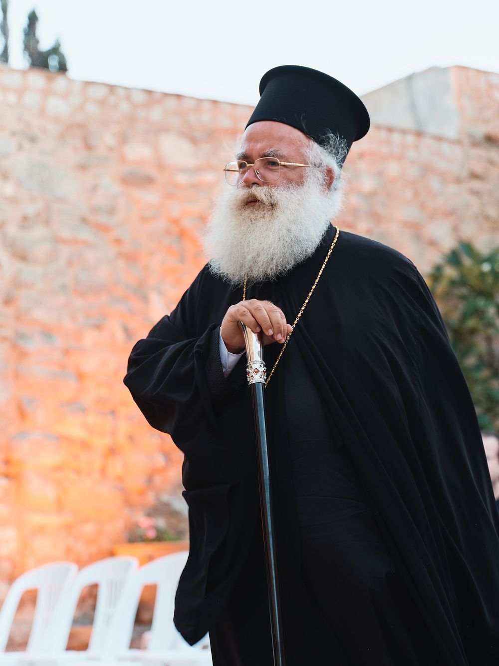 Orthodox clergy member in traditional black robes photographed against sunset-lit stone wall.