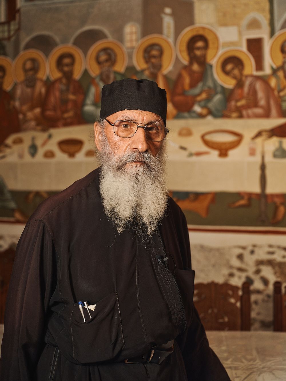 Orthodox religious figure seated inside church with traditional Byzantine murals in background.