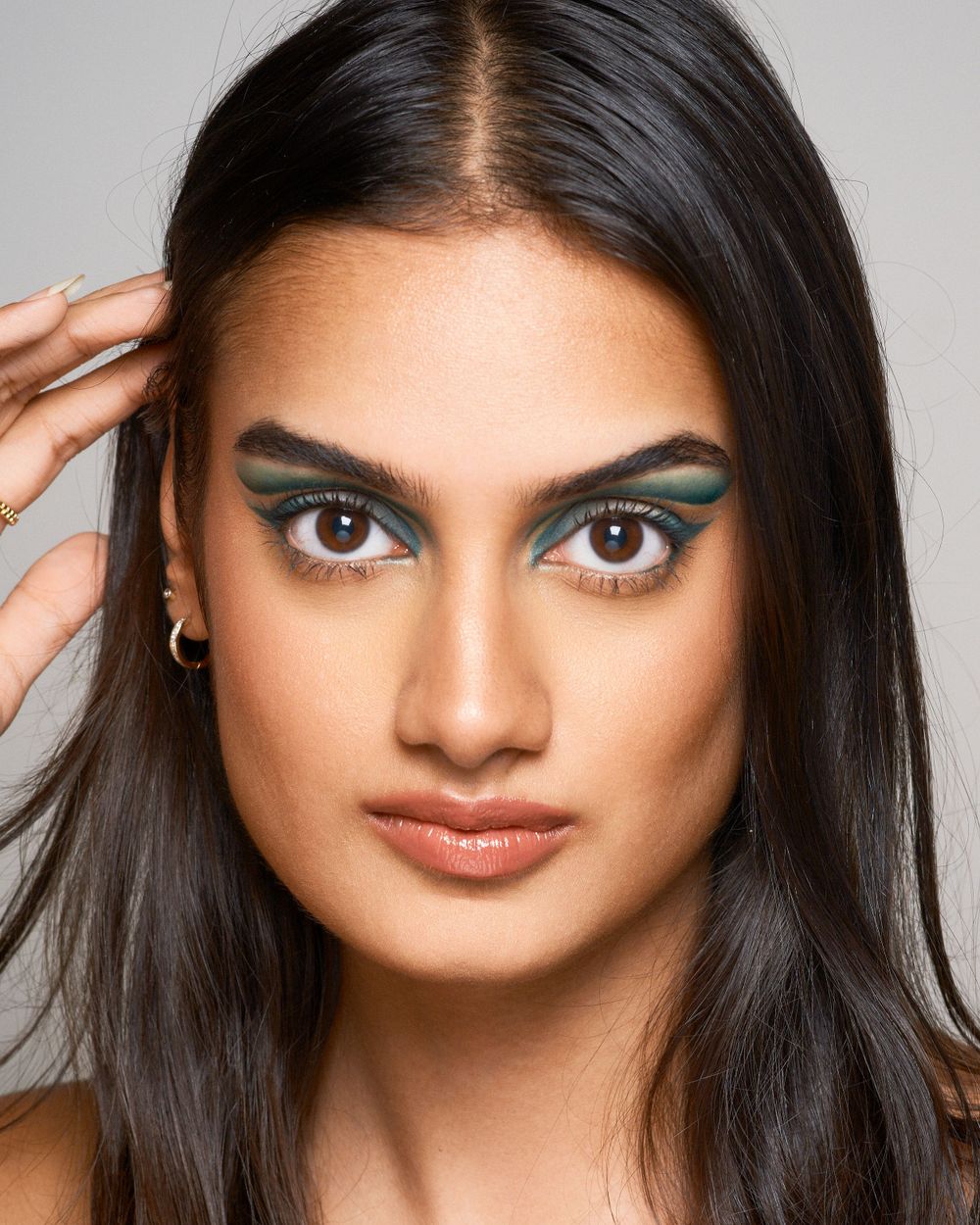 Close up beauty portrait showing artistic teal eyeshadow and styled dark hair against light background.