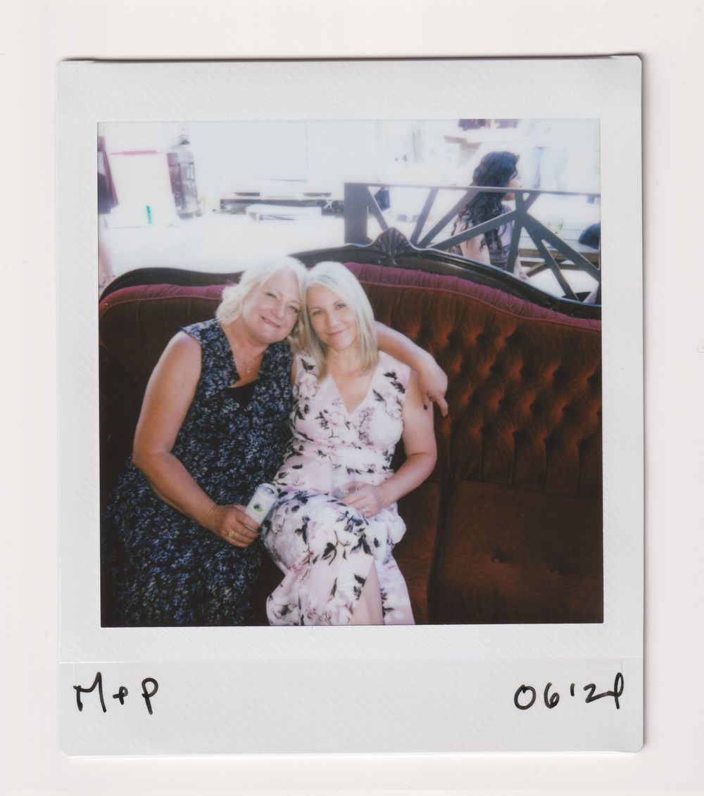 Two people in floral dresses sit together on a brown leather couch in a casual setting.