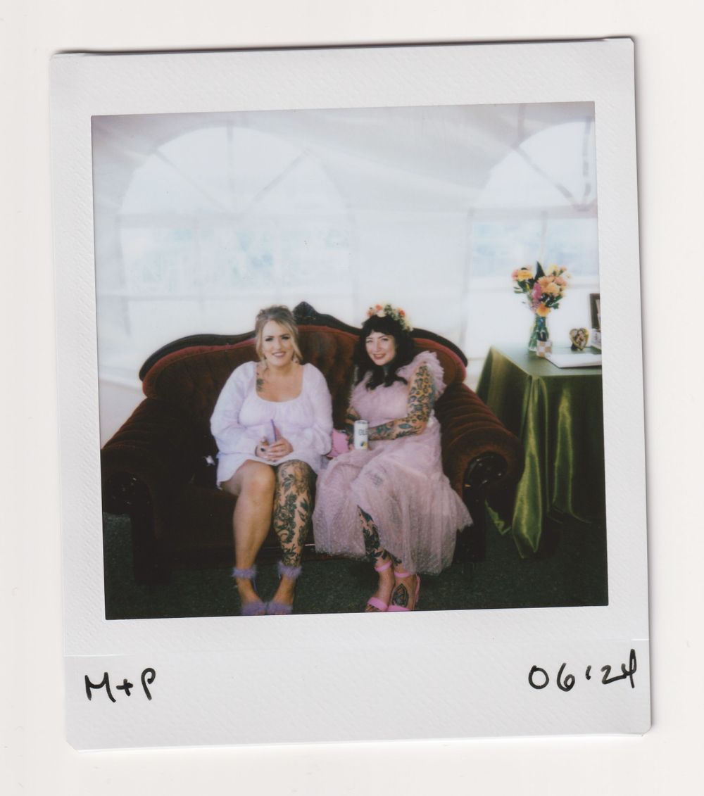Two people in light-colored dresses sitting on a vintage brown sofa with floral arrangement nearby.