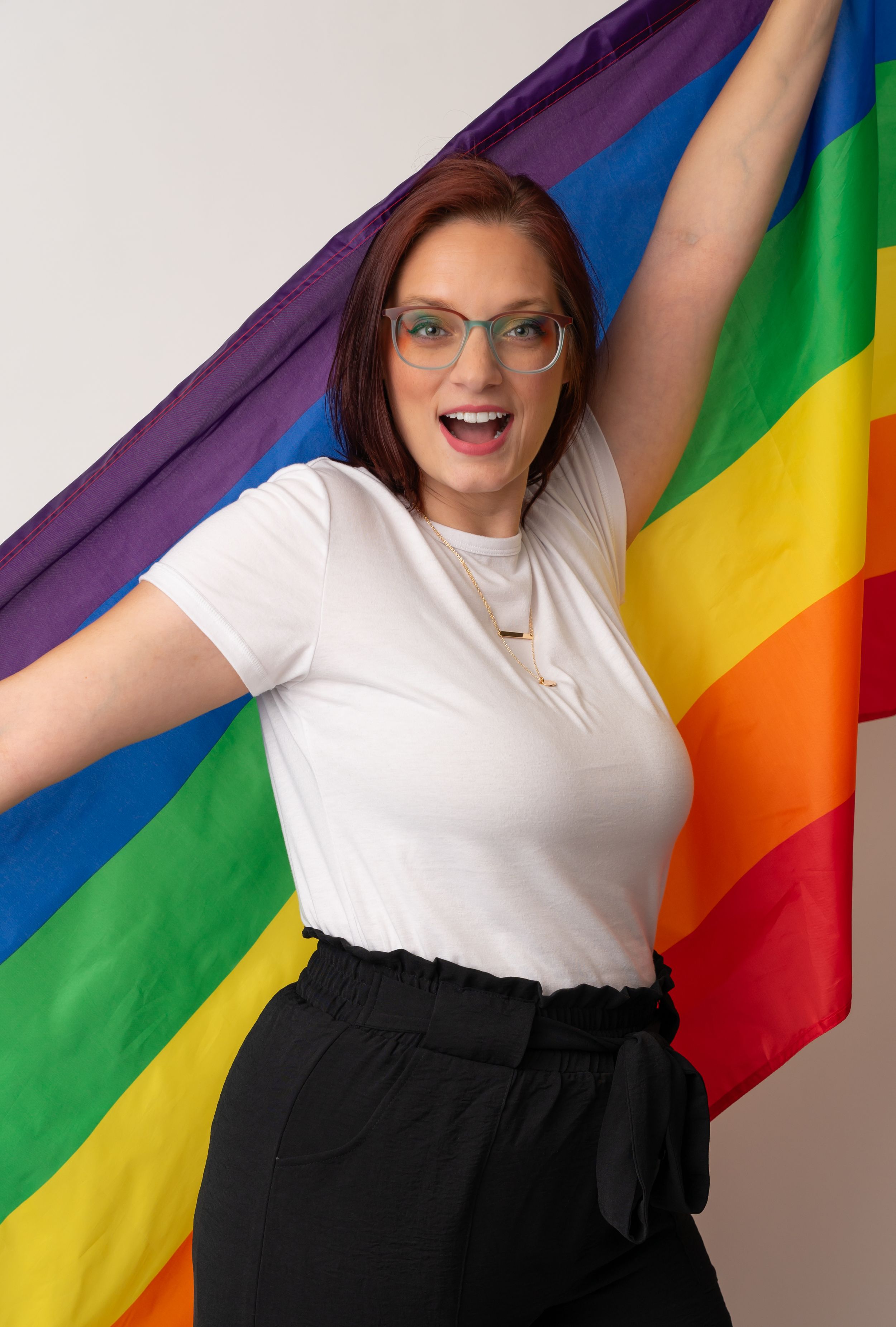 A person wearing glasses poses joyfully with arms spread wide against a Pride flag backdrop.