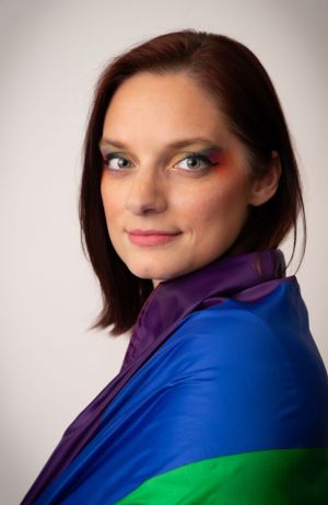 A portrait featuring someone wrapped in a vibrant rainbow Pride flag against a neutral background.