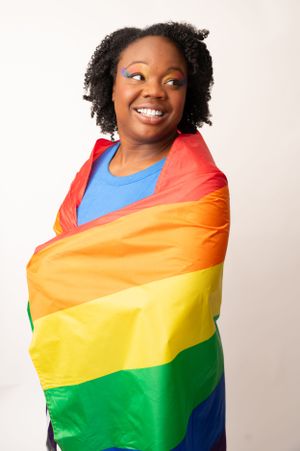 A person wrapped in a Pride flag smiles warmly against a white studio background.