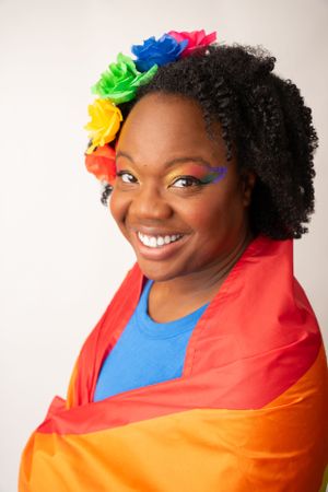 A person wearing rainbow flowers and Pride flag displays colorful makeup against white background.