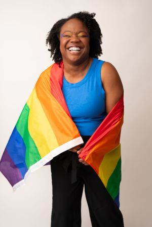 A person in blue poses joyfully while wrapped in a rainbow Pride flag.