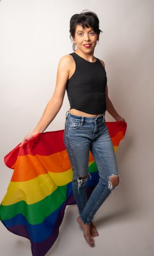 Person in black top and jeans poses energetically with rainbow pride flag.
