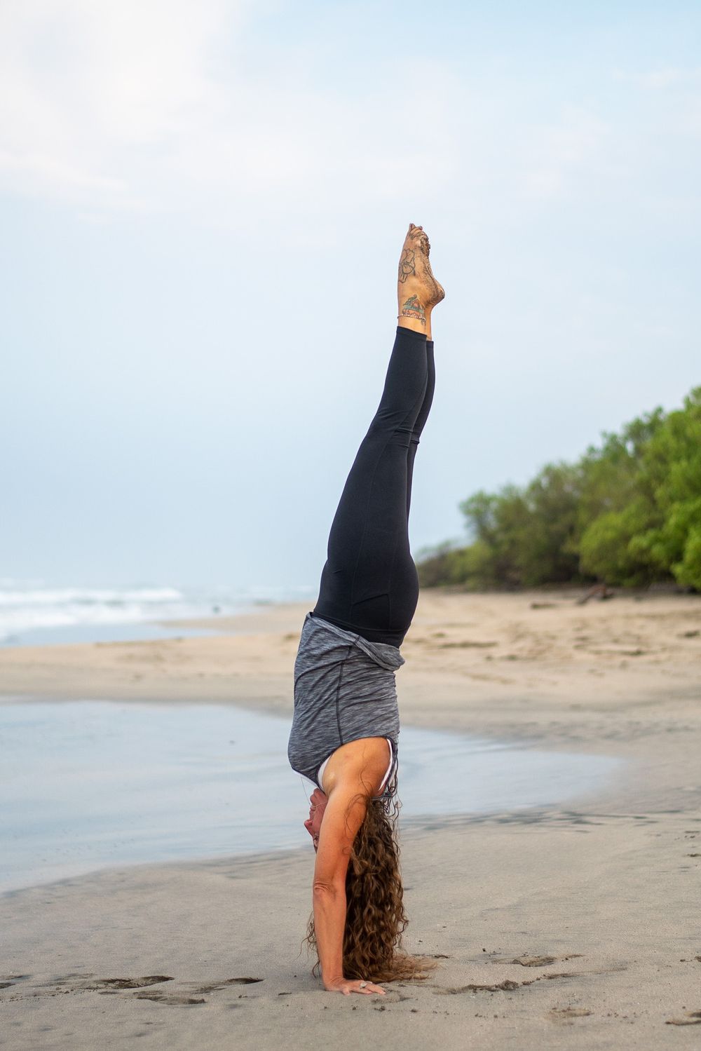 Person performing handstand on beach with ocean and trees in background.
