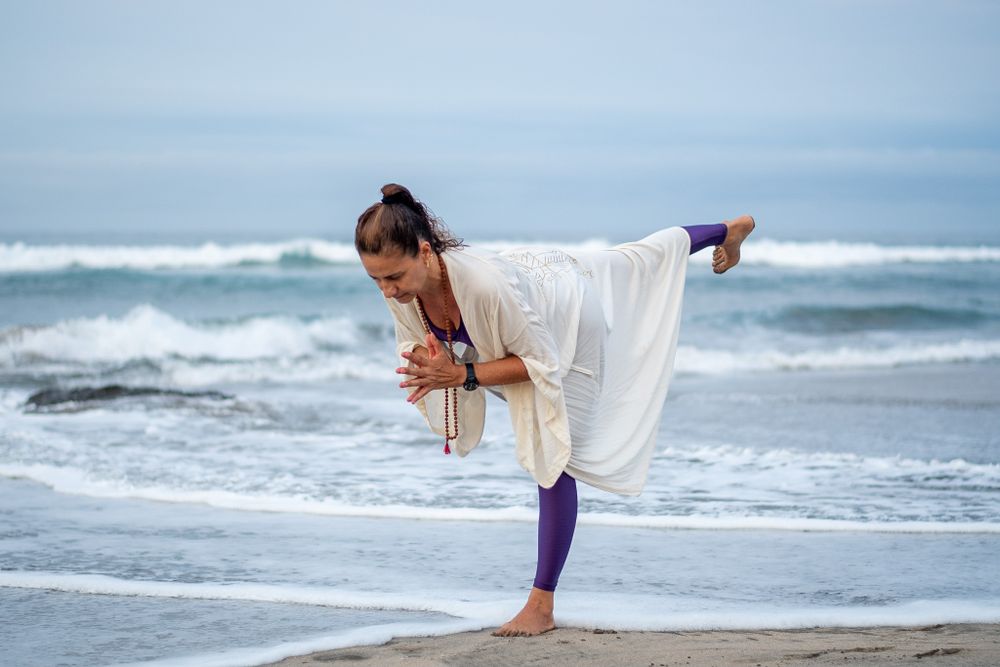 Person in white flowing dress performing graceful yoga pose on beach with waves in background.