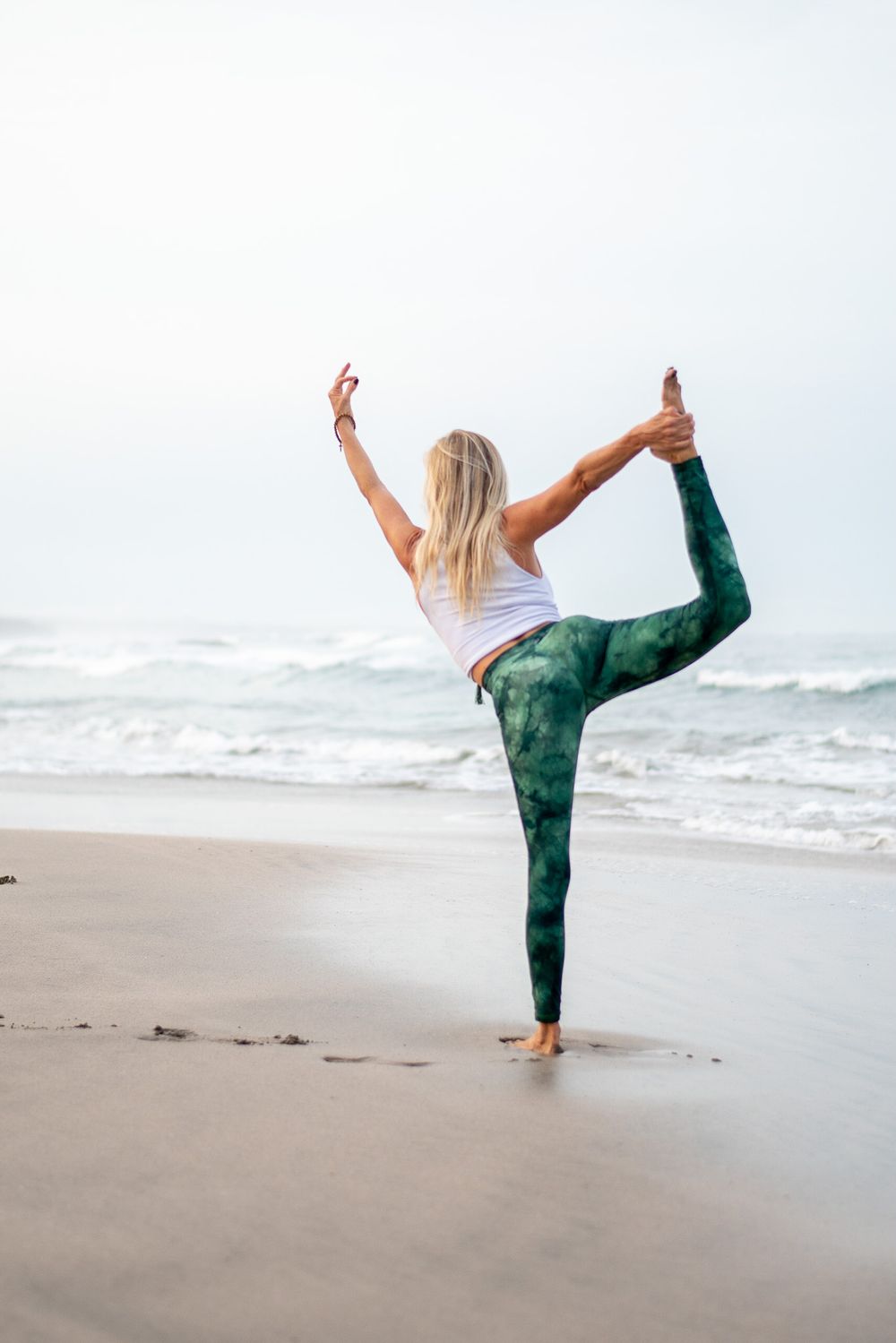 Yoga practitioner performs dancer pose on beach in green patterned leggings and white tank top.
