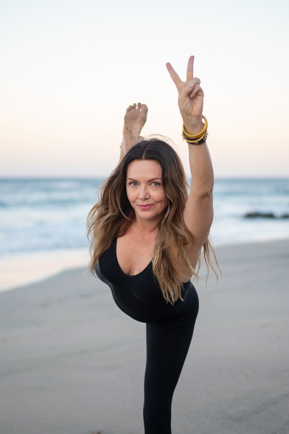 Person in black yoga outfit demonstrating standing split pose on beach at dusk.