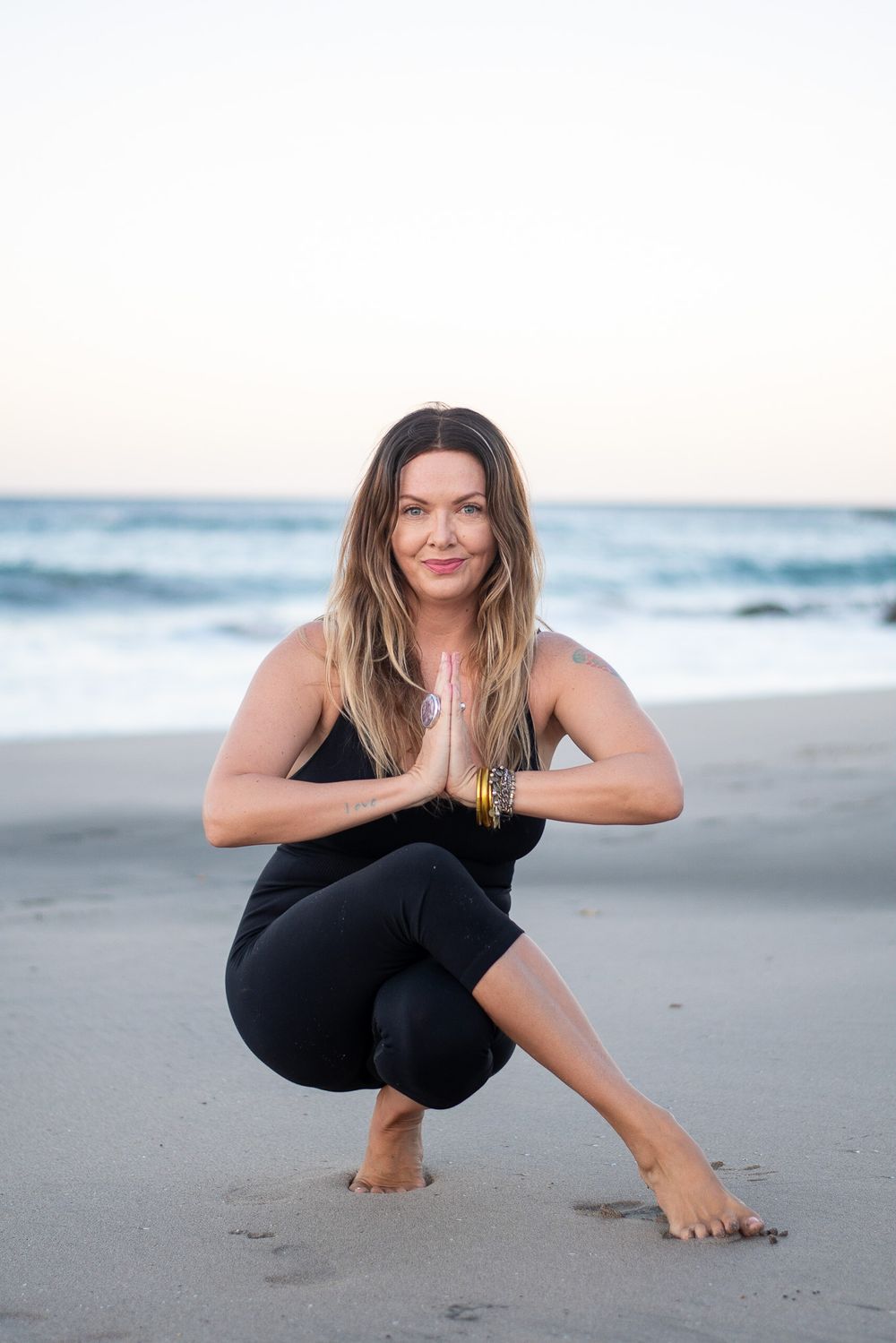 Person in black athletic wear demonstrating squat yoga pose on sandy beach.