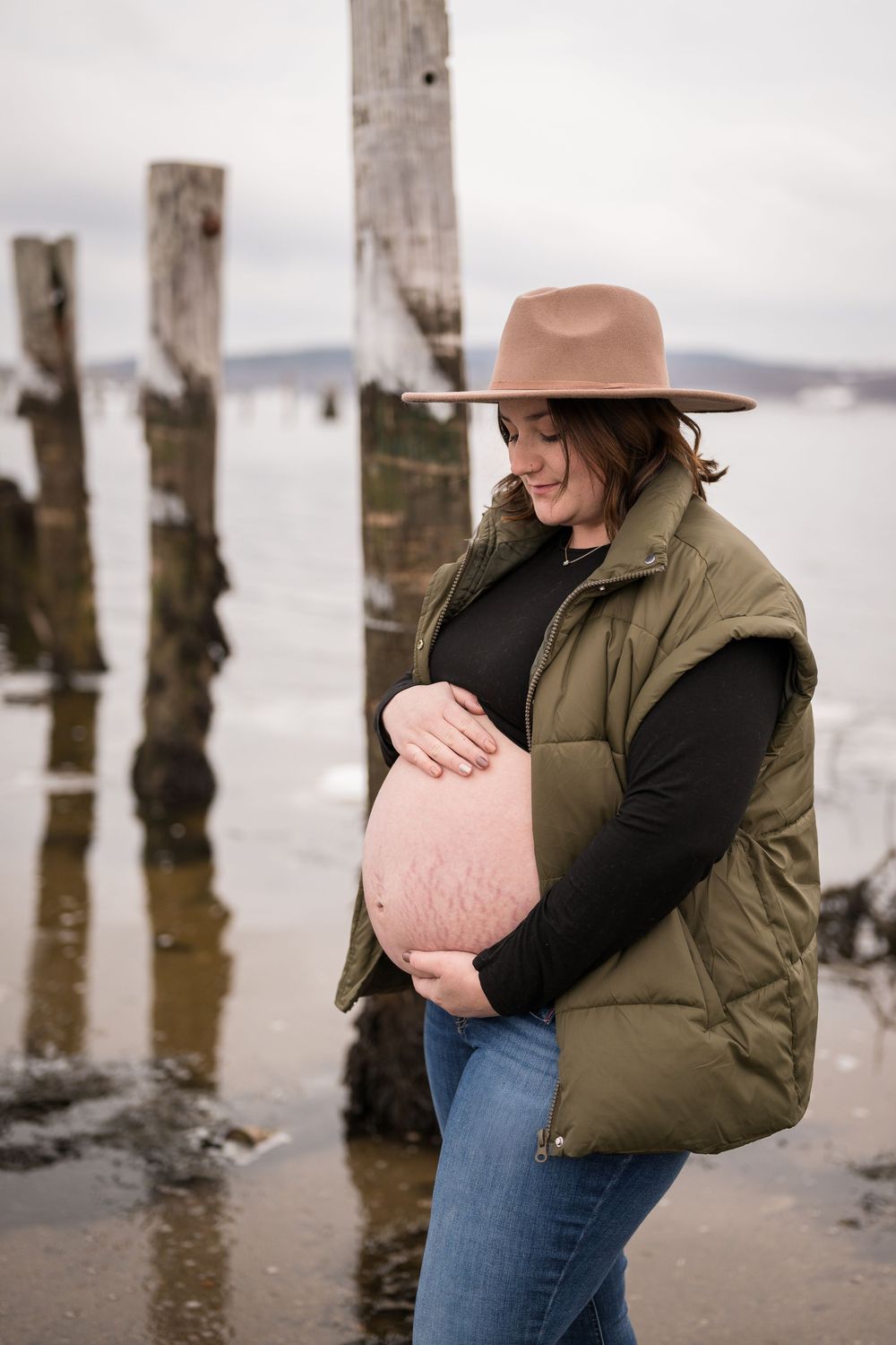 A person in a brown jacket and jeans stands near wooden posts by the water's edge in a serene coastal setting.
