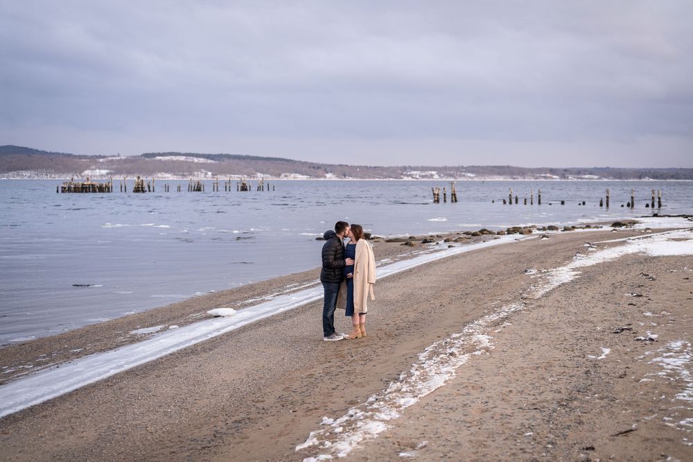 Couple walking along a snowy winter beach with waves crashing on the shore at dusk.