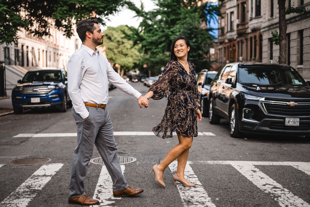 A playful engagement photo session on a city crosswalk with parked cars in the background.