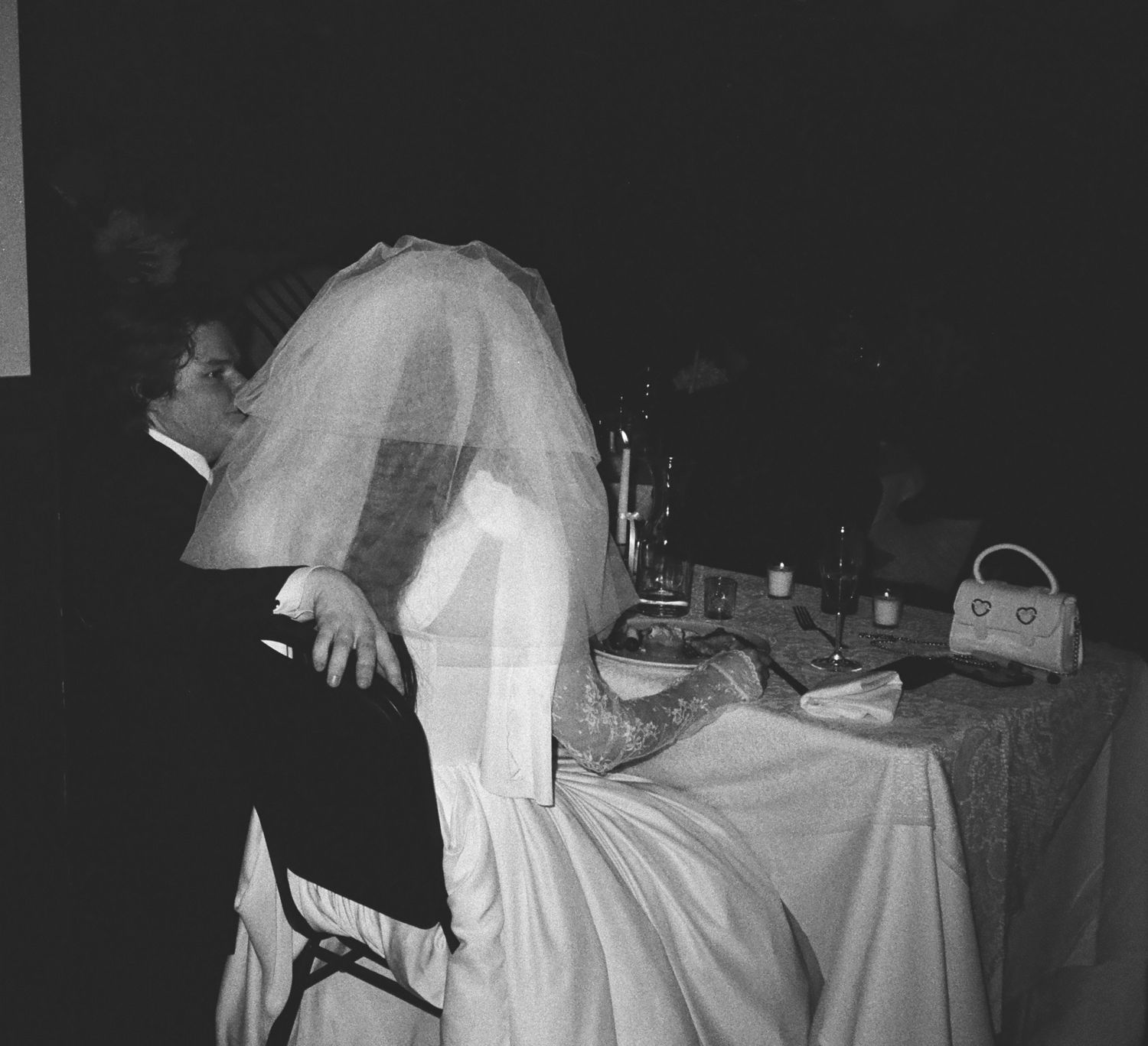 Woman in white wedding dress seated at table with dark silhouette beside her.