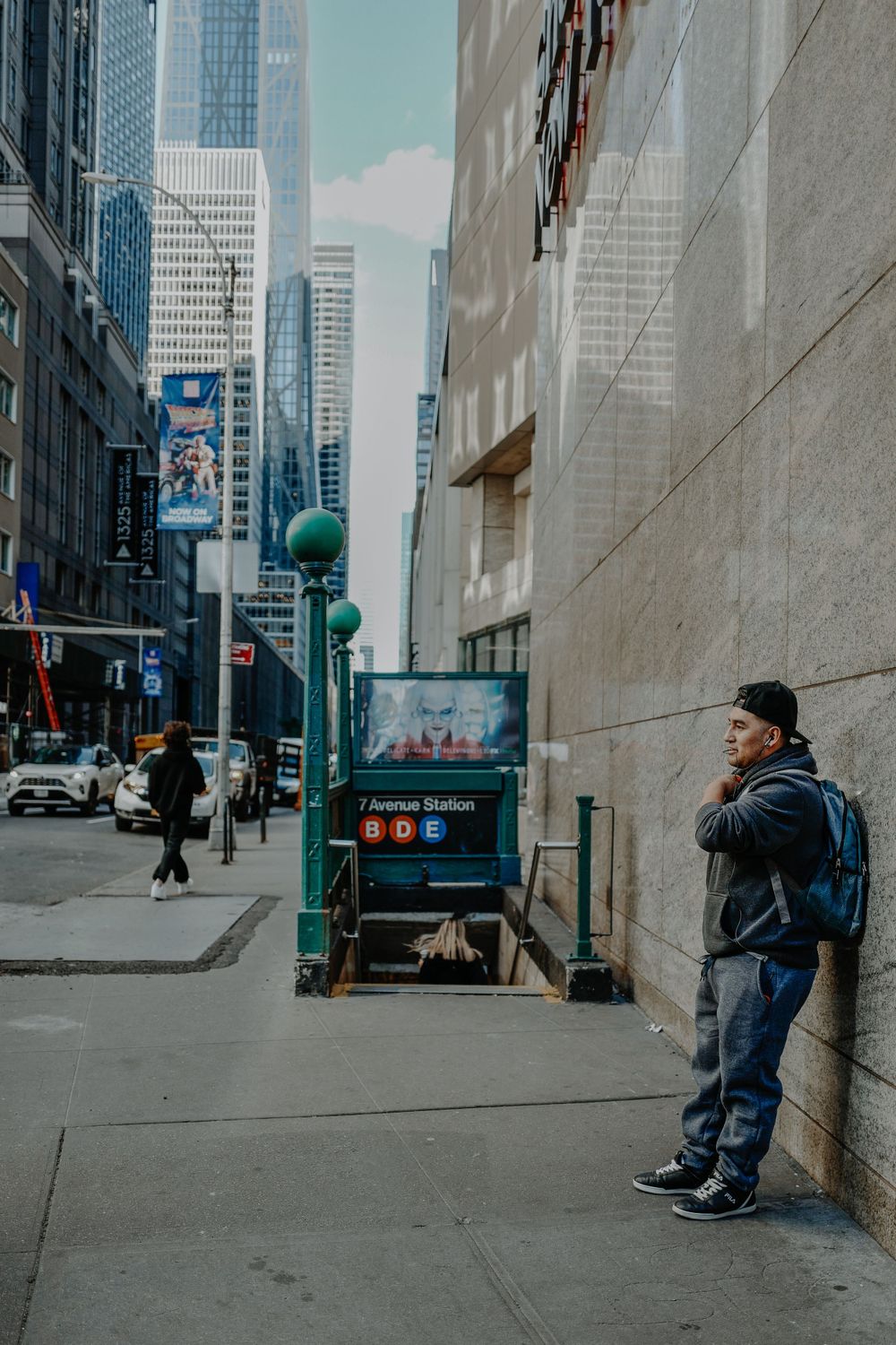 Person with backpack stands against wall on urban street with tall skyscrapers in background.