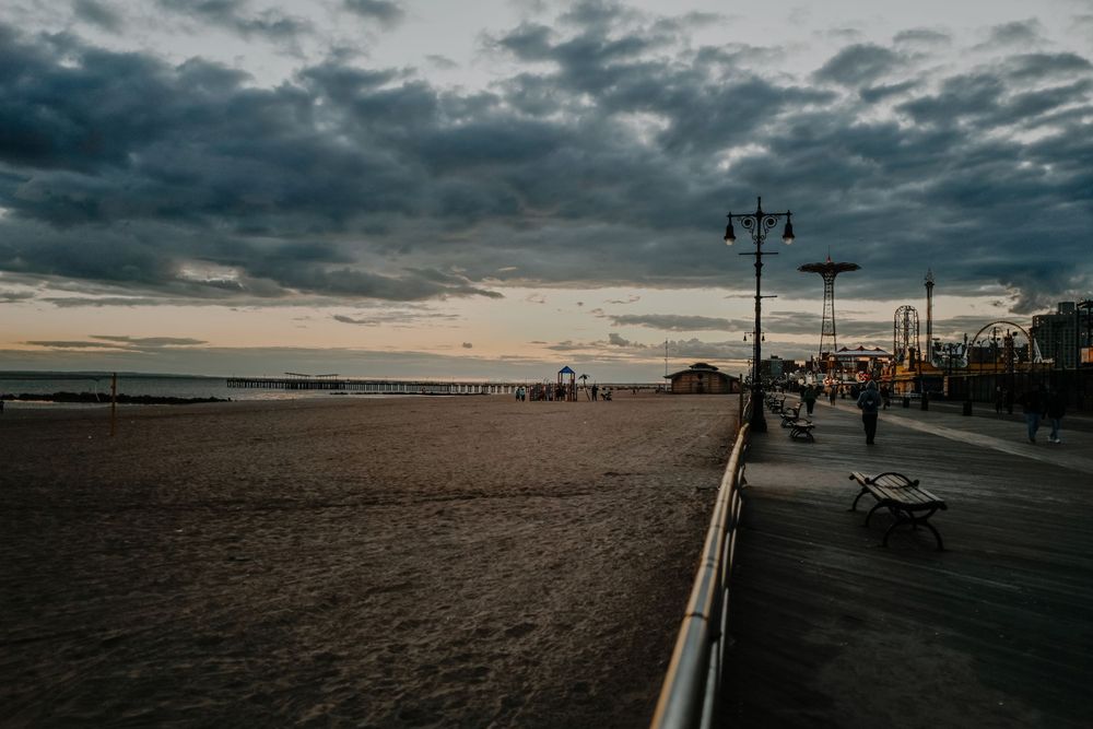 A dramatic sunset over a beach boardwalk with storm clouds and lampposts silhouetted against the sky.