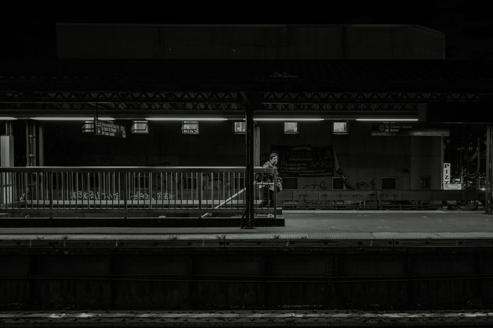 A dark and moody black and white photograph of an empty train platform at night.
