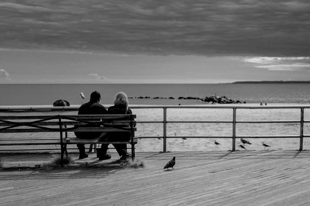 Three people sit on boardwalk bench overlooking ocean in black and white photograph.