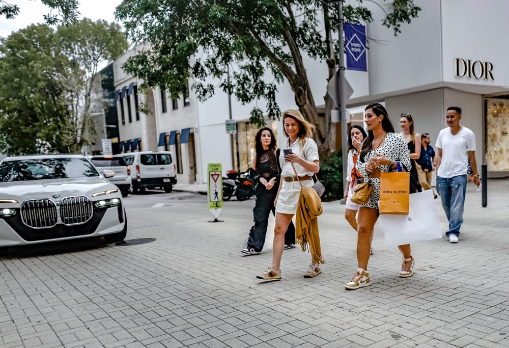 Shoppers walking past luxury stores and parked cars on an upscale urban street.