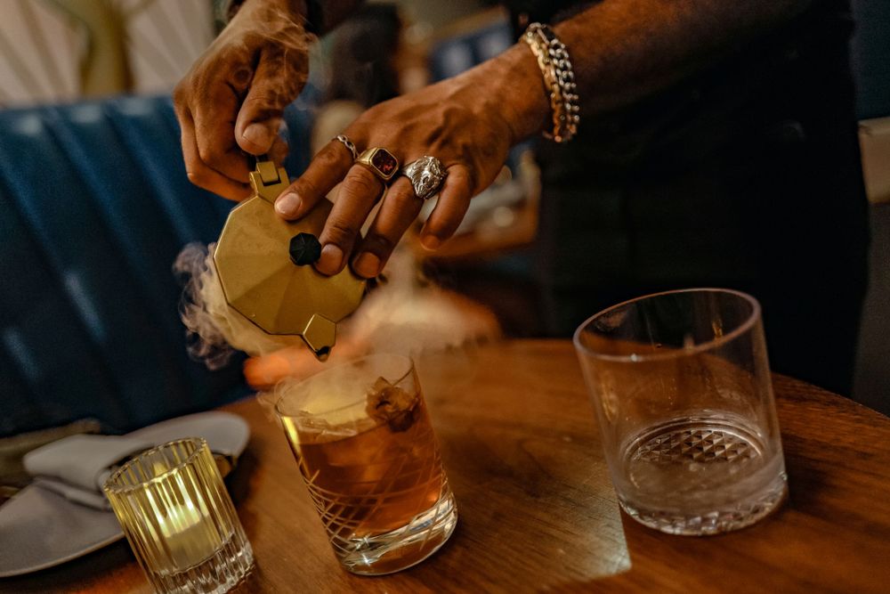 Hands pouring a cocktail at a dimly lit bar with glowing candles and glasses on wooden table.