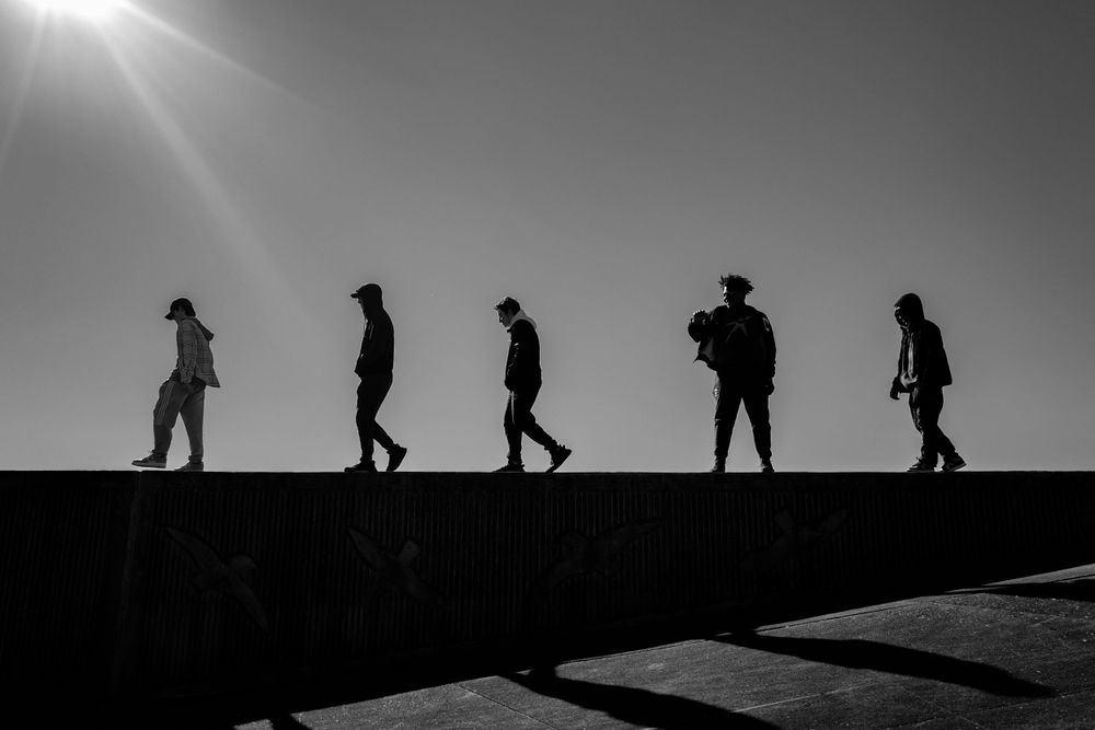 Black and white silhouettes of people walking along a ridge against bright sunlight.