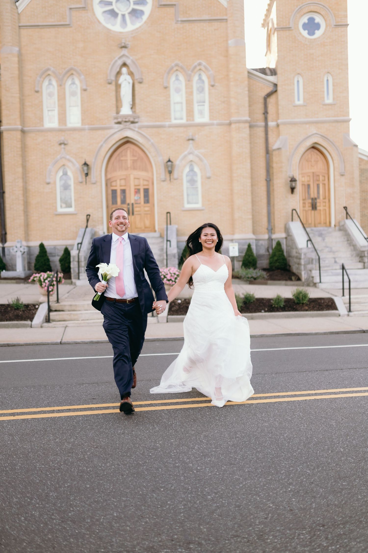 Newlyweds walk hand-in-hand across a street in front of a Gothic stone church, bride in white gown, groom in navy suit.