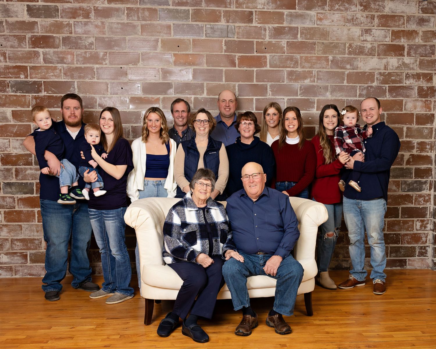 A large multigenerational family poses together in front of a brick wall, with an elderly couple seated in the center.