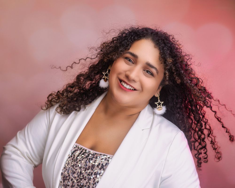 Smiling woman with curly hair in a white blazer and star earrings, standing against a soft pink bokeh background Image Carolina Waldroff