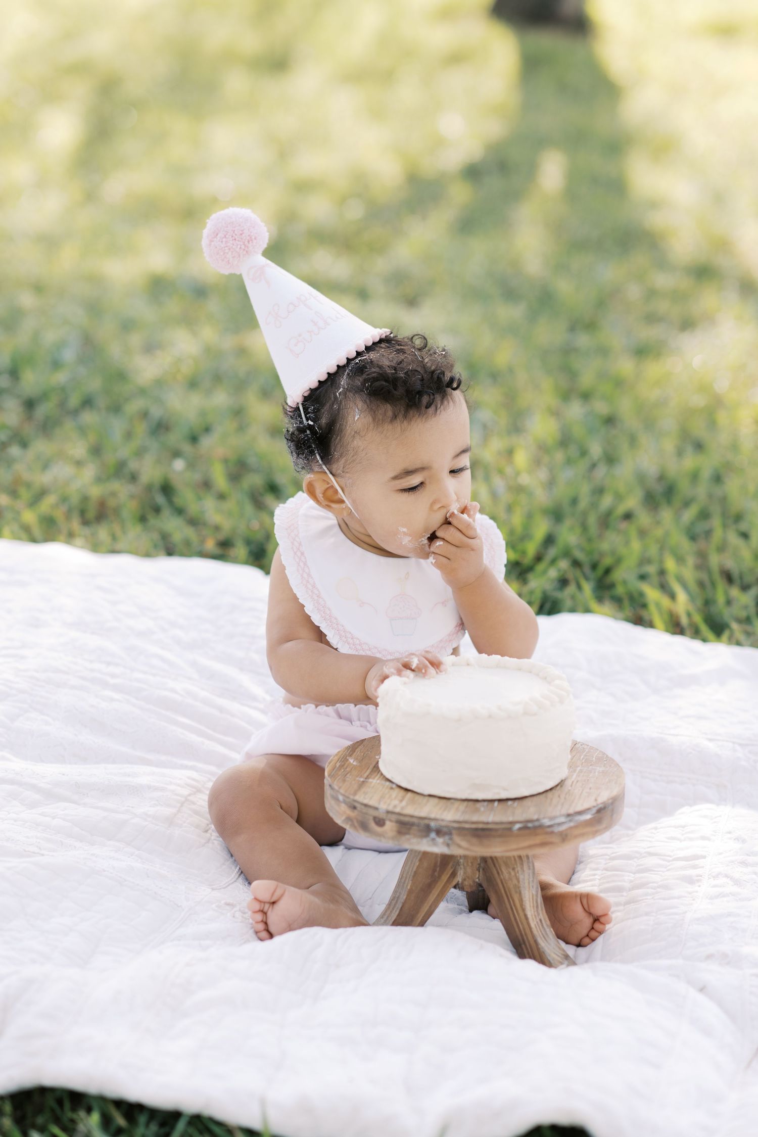 A baby in a pink party hat explores a white smash cake on a blanket in a sunny outdoor setting.