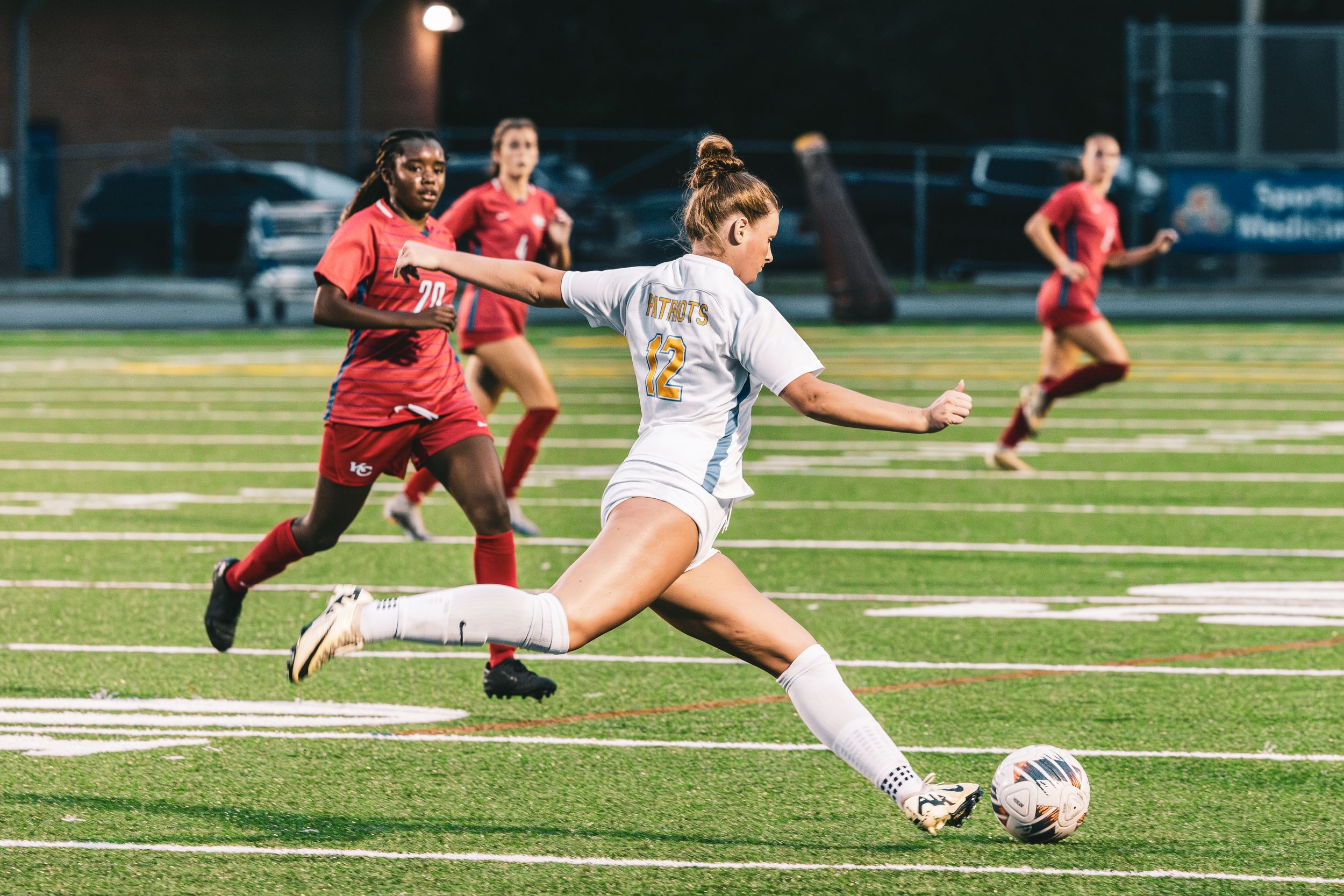 Female soccer player in white uniform kicks ball during evening match.