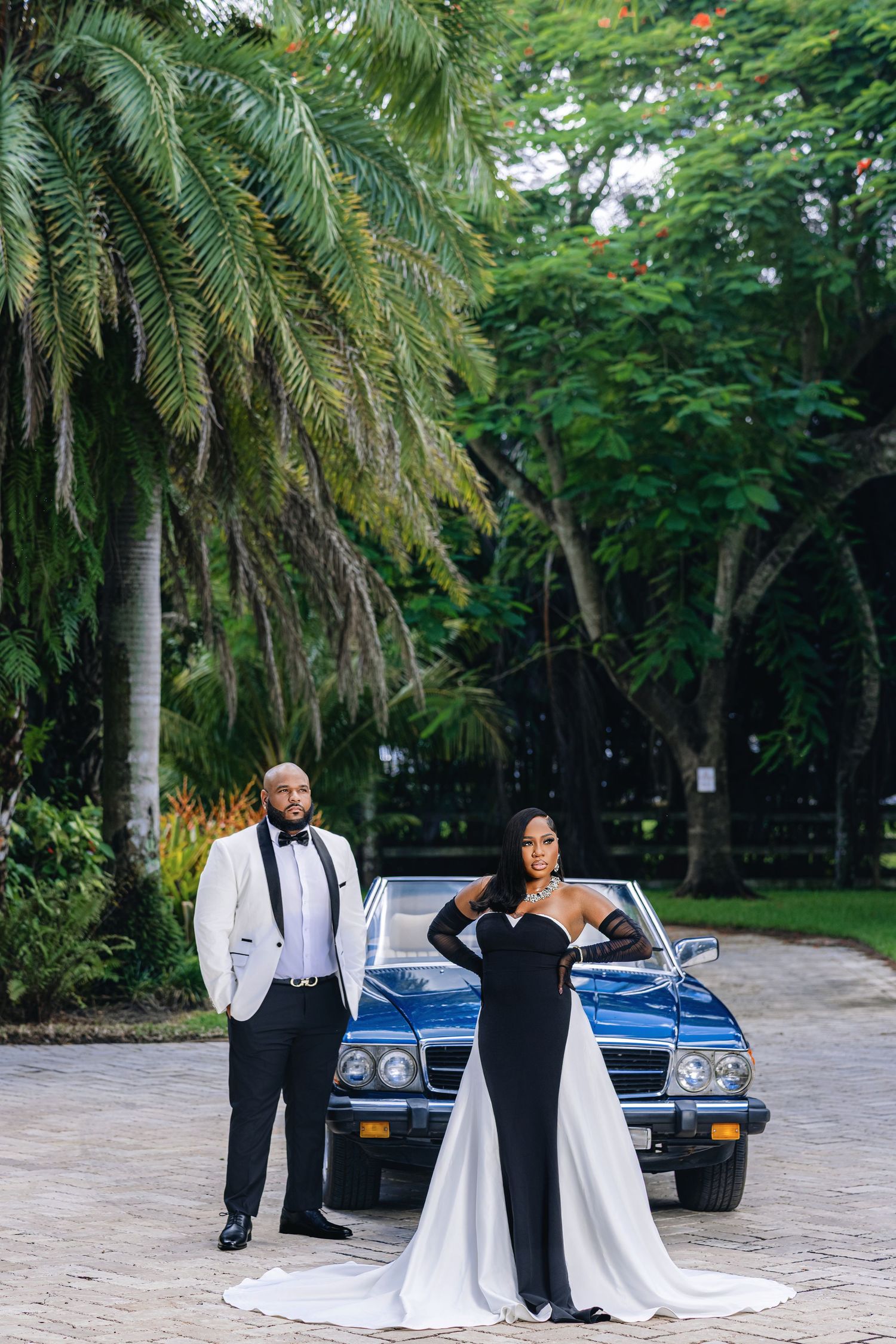 Elegant couple in black and white formal wear poses in front of a vintage blue convertible outdoors.