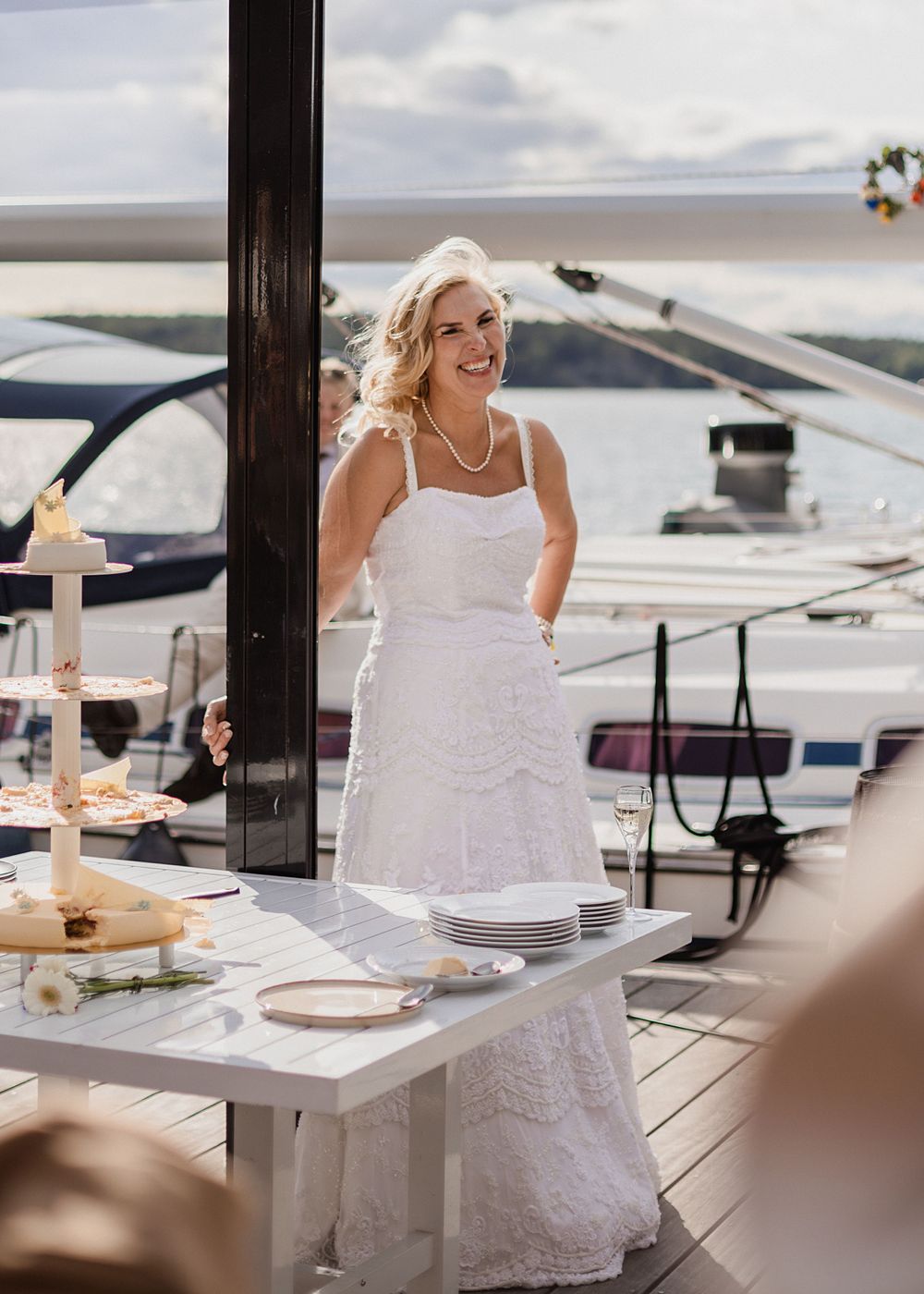 Smiling bride in white lace wedding dress standing on a yacht deck with a tiered cake nearby.