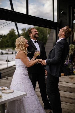 Bride in white lace gown laughing with two suited guests on a waterfront deck during wedding celebration.