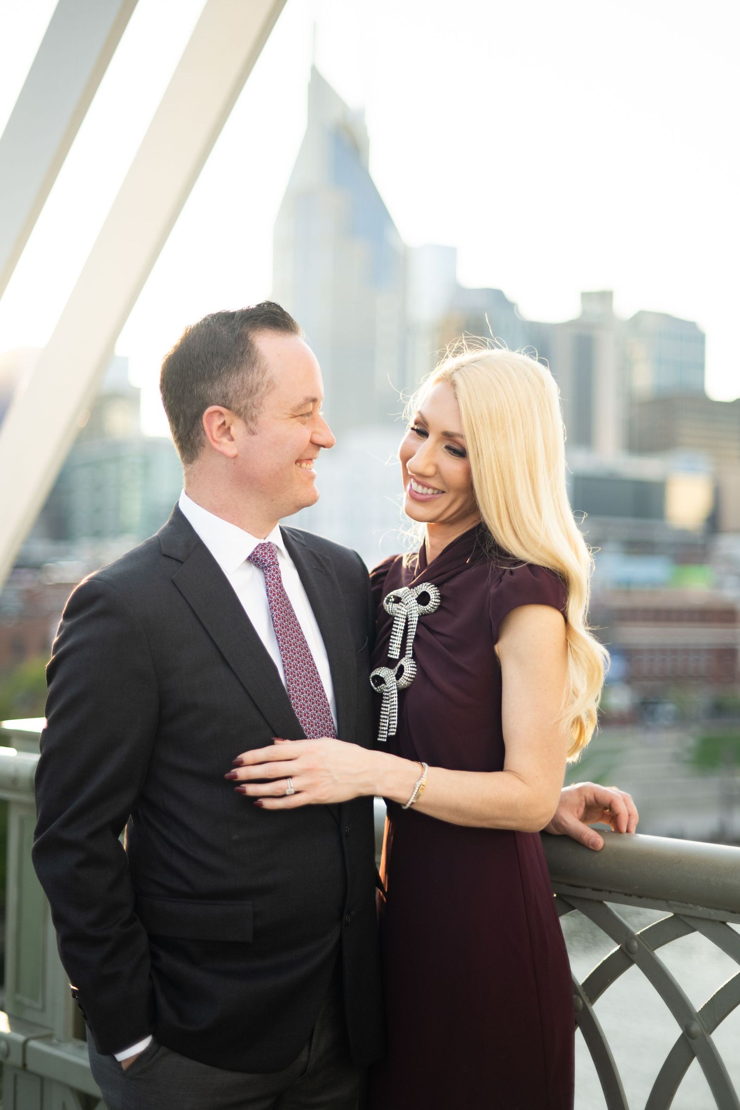 Elegant couple sharing a romantic moment at sunset, with her in burgundy and him in a dark suit on Pedestrian Bridge in Nashville.