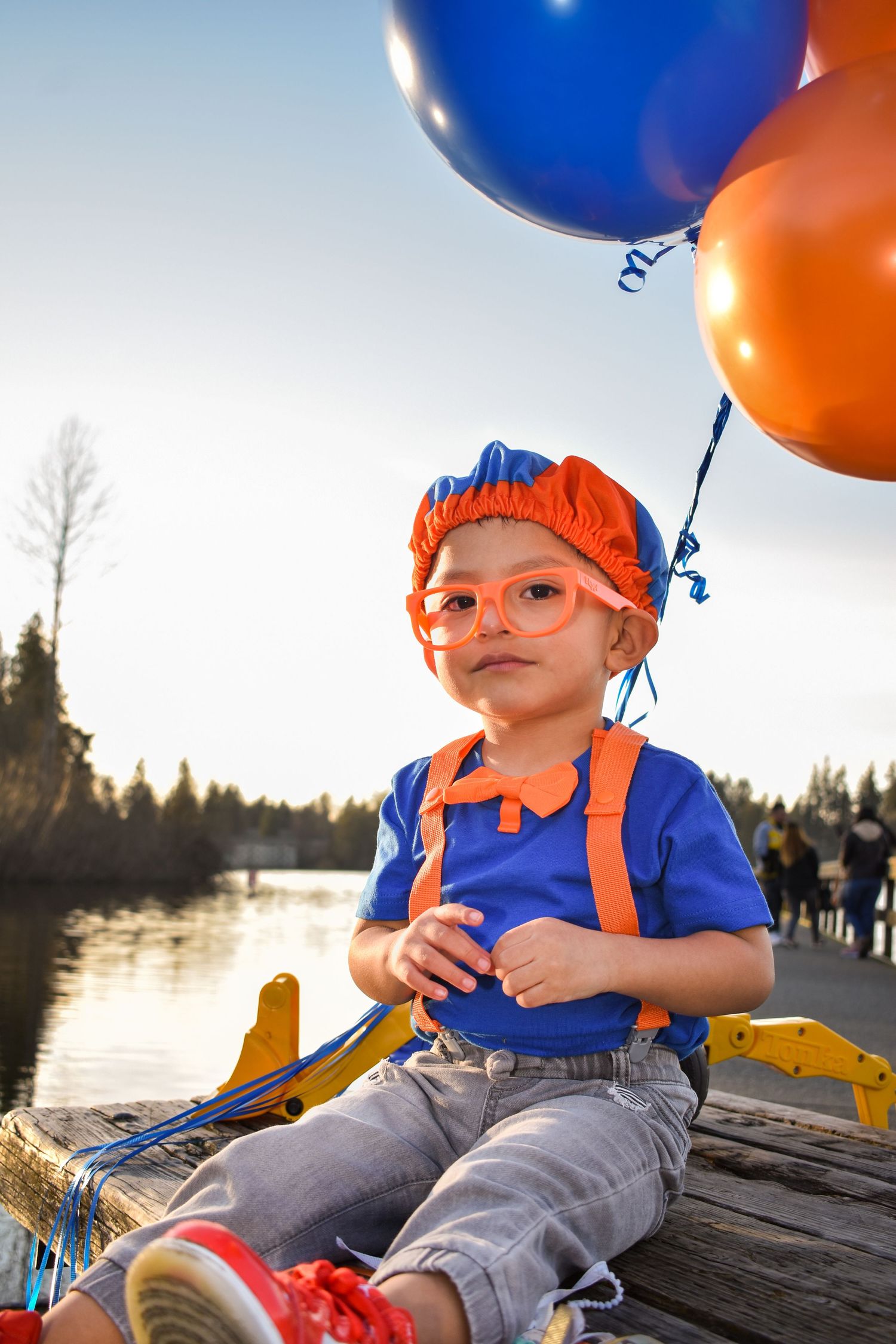 Child in orange hat and overalls holding colorful balloons by lakeside at sunset.