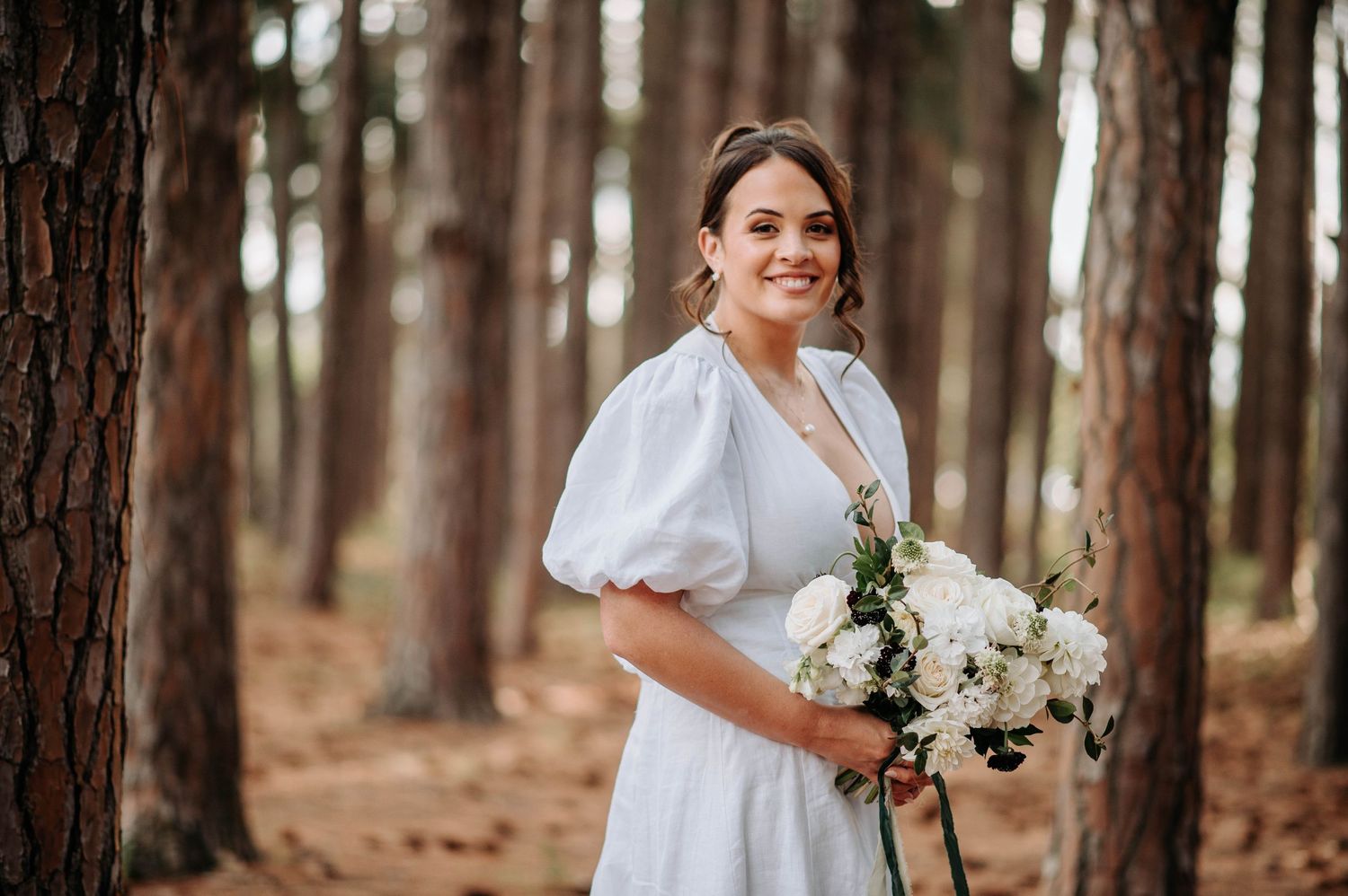 A bride in a white dress holding a bouquet of white flowers in a pine forest.
