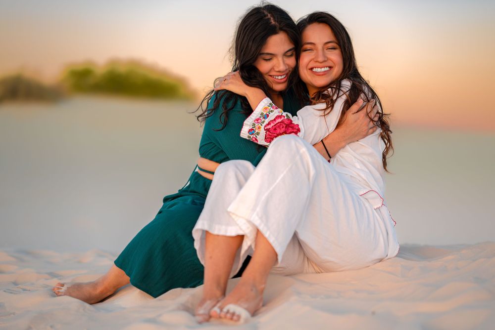 Photograph of Two friends embracing on beach sand during sunset.