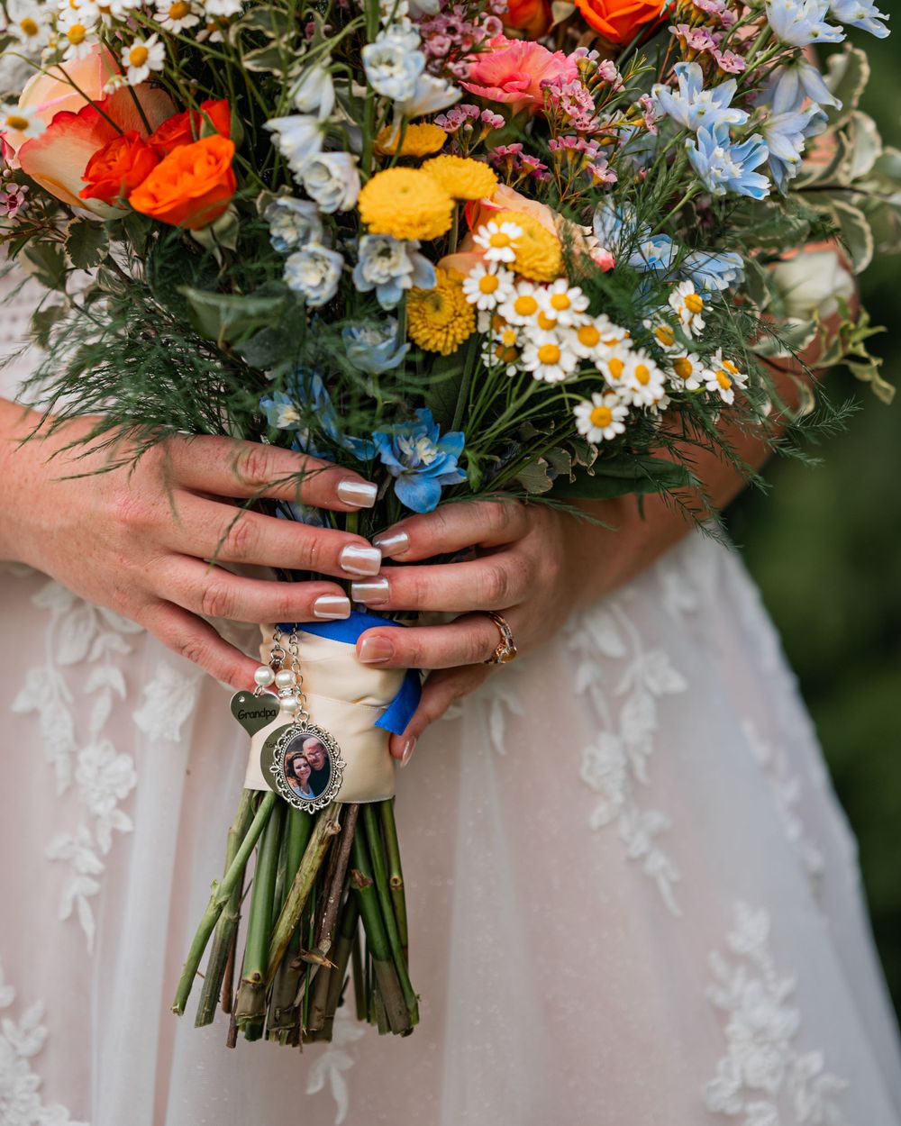Colorful wedding bouquet with blue ribbon and vintage brooch held by hands in white dress.