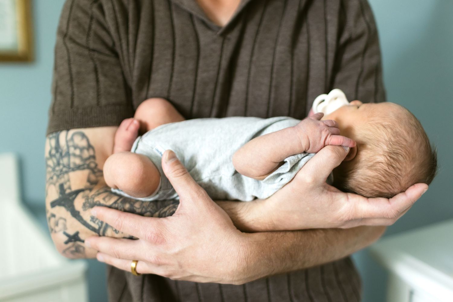A father with a cross tattoo wears a brown short sleeved sweater holding his infant son wearing a gray onesie as he sucks a pacifier.