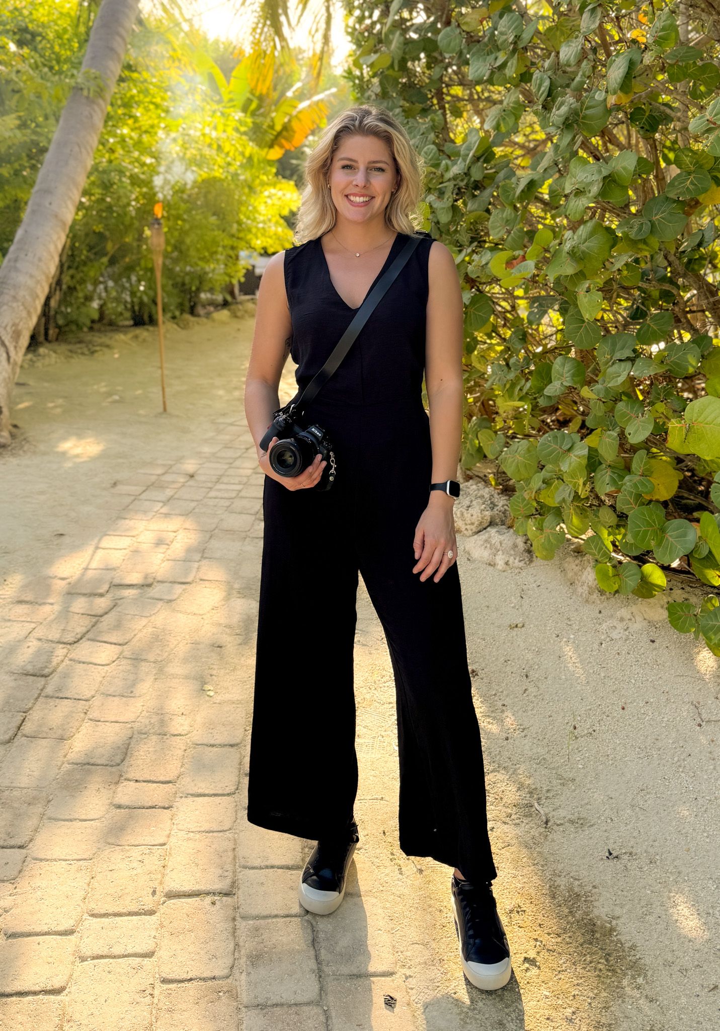 A photographer in a black jumpsuit stands on a brick path surrounded by lush greenery and golden sunlight.