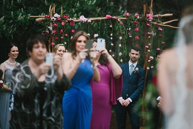 Wedding guests taking photos with phones during outdoor ceremony under floral arch.