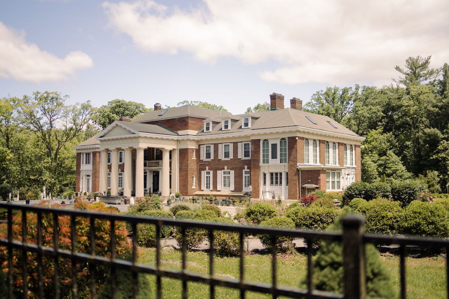A grand red brick colonial mansion with white columns sits surrounded by mature trees under a partly cloudy sky.