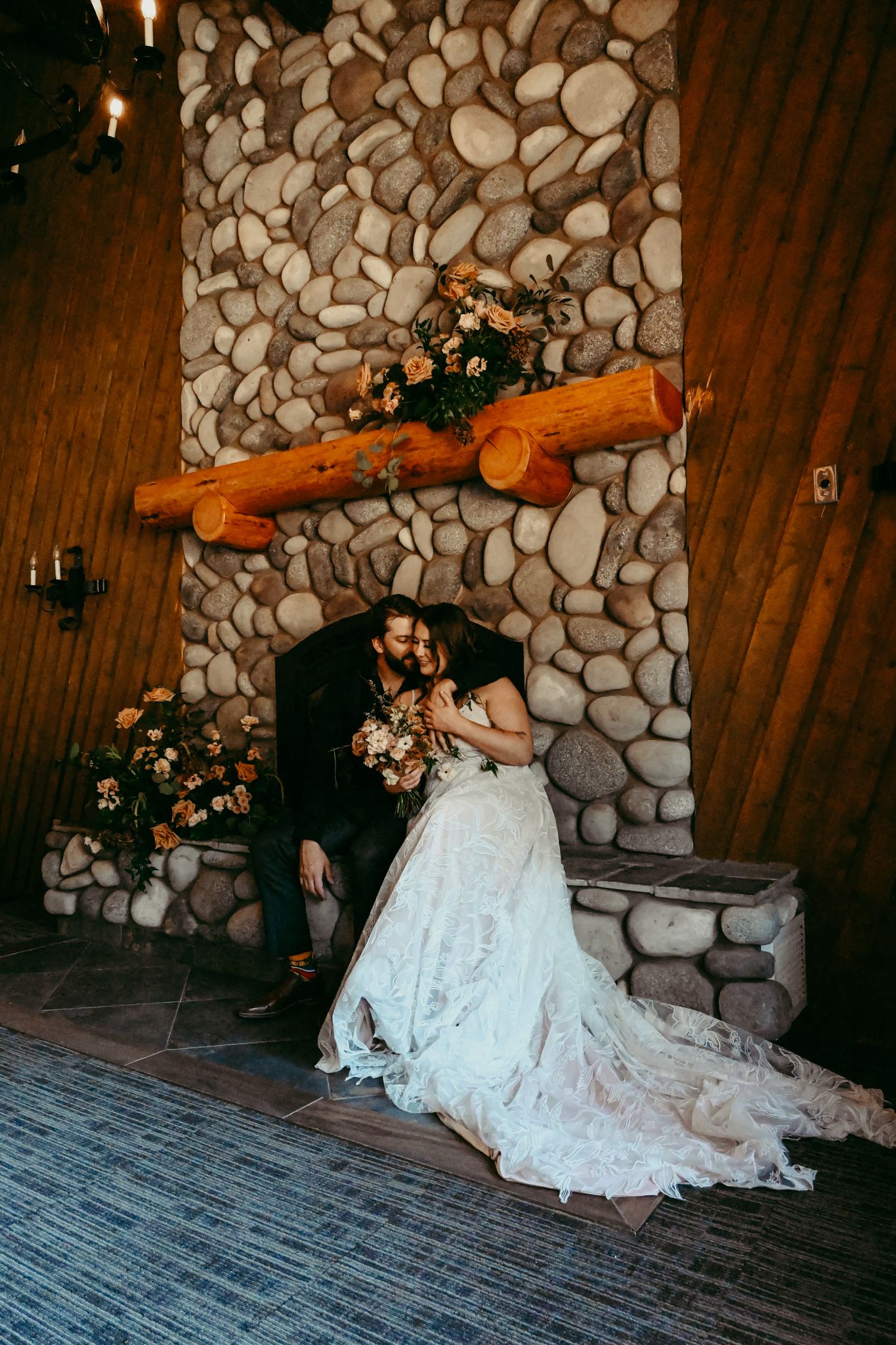 Newlyweds sharing intimate moment by stone fireplace in rustic indoor setting.