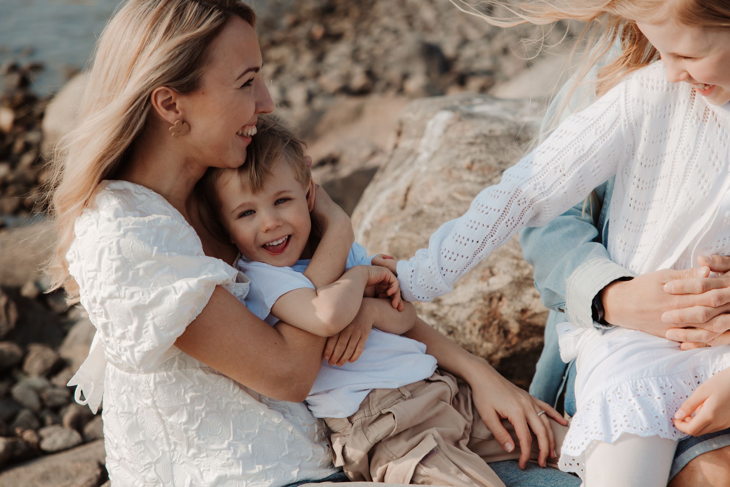 Family in white outfits sharing tender moment on rocky beach.