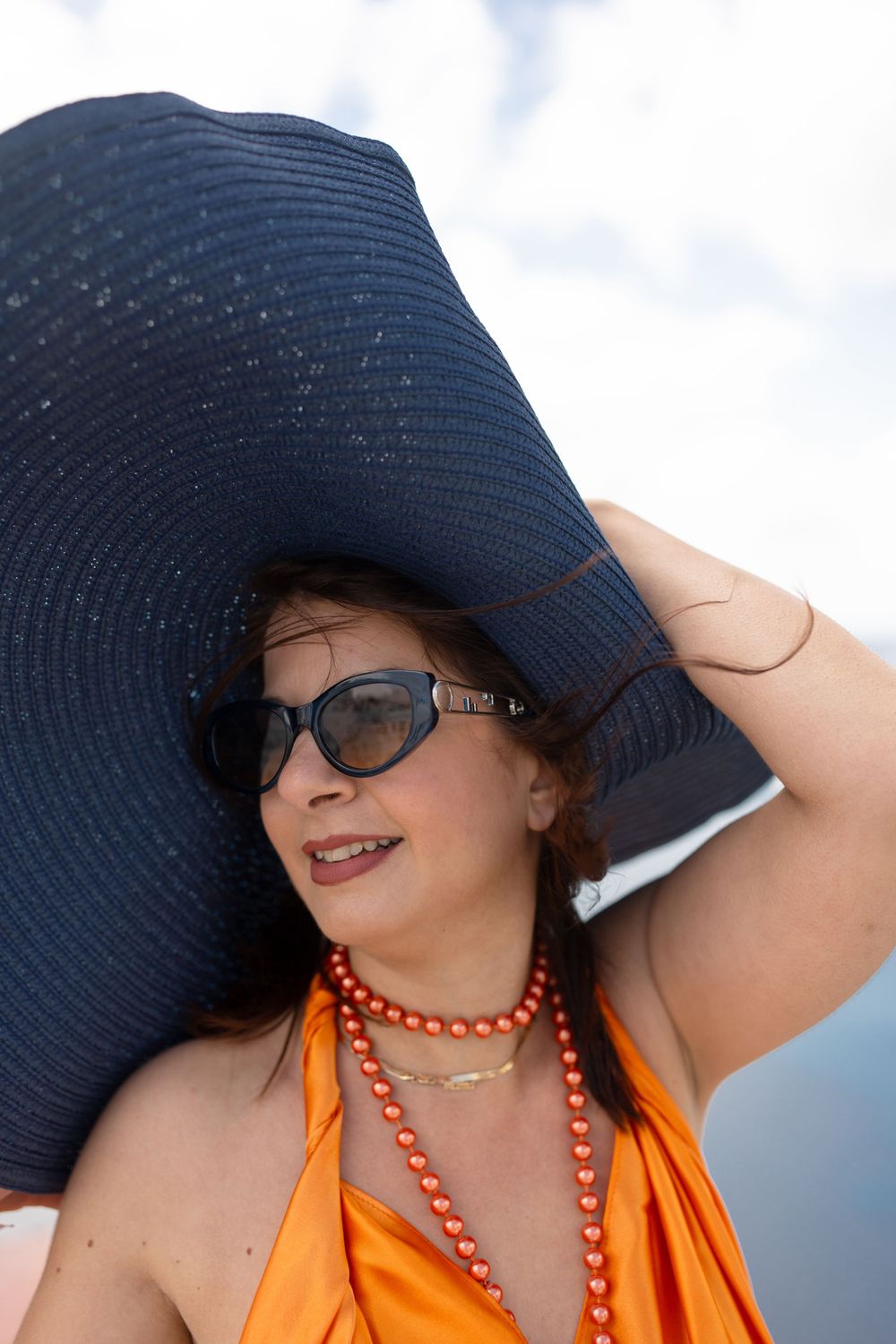 Summer fashion with large navy sun hat and orange dress against cloudy sky backdrop.