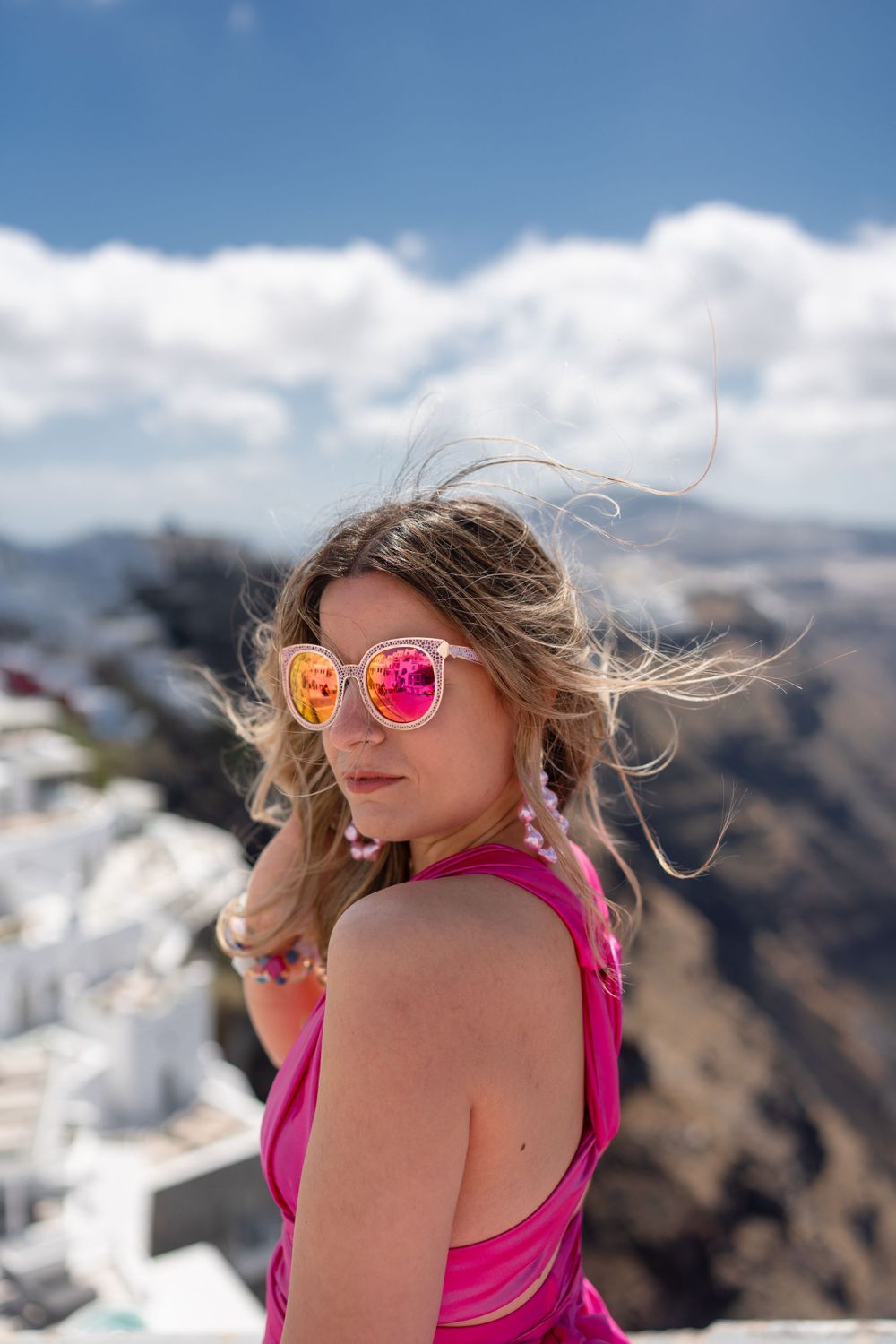 Pink-dressed figure with mirrored sunglasses against Santorini's white architecture.