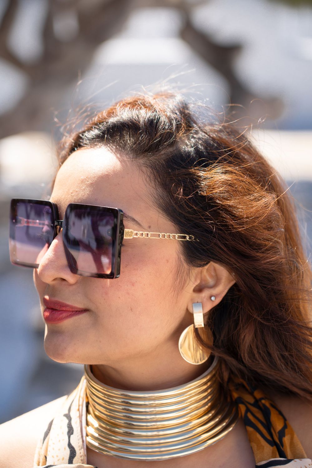 Close-up of statement gold choker necklace and oversized sunglasses against coastal background.